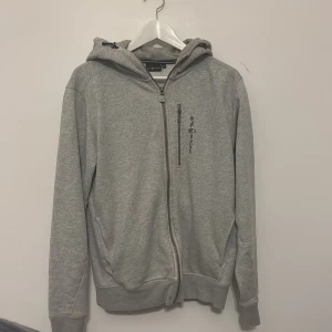 Grå hoodie från Sail Racing M - Grå hoodie från Sail Racing med dragkedja och huva. Snyggt broderad logga på bröstet och praktisk bröstficka med dragkedja. Mjuk och skön bomullsblandning, perfekt för chill dagar. Justerbar huva med snörning och ribbade muddar.