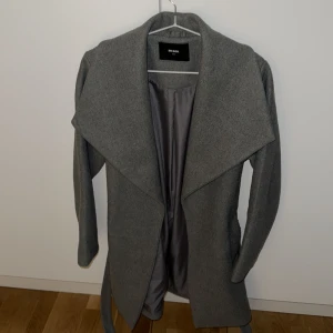 Grå kappa med bälte från Bik Bok XS - Stilren grå kappa från Bik Bok i storlek XS. Jackan har bred krage, knytskärp i midjan och raka fickor. Perfekt för höst och vår, tillverkad i mjukt material med slät insida. Enkel och snygg design som passar till det mesta.