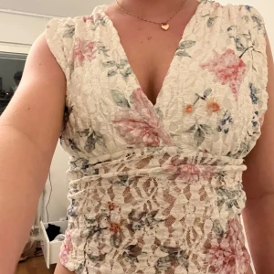 Blommig spetstopp från Gina Tricot - Supersöt topp från Gina Tricot i vit spets med blommigt mönster i rosa, blått och grönt. Djup v-ringning och omlottkänsla upptill, ärmlös design och figurnära passform.