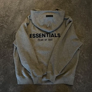 Grå Essentials Fear of God hoodie M - Säljer en grå hoodie från Essentials Fear of God i storlek M. Tröjan har svart tryck på bröstet, huva och ribbade muddar. Materialet är mjuk bomullsmix med 80% bomull och 20% polyester. Perfekt för en chill och stilren look.