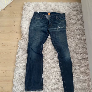 Blå jeans från Boss Orange - Snygga blå jeans från Boss Orange med slitningar och rivet hål på ena låret. Klassisk femficksmodell med raka ben och kontrastsömmar. Jeansen har en mörk tvätt och är tillverkade i mjukt bomullsjeansmaterial.