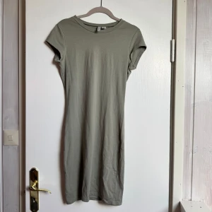 Grön kortärmad klänning från H&M - En enkel och stilren grön klänning från H&M med rund hals och korta ärmar. Modellen är figurnära och har en rak siluett som slutar strax ovanför knäna. Tillverkad i mjuk bomull som känns skön mot huden. Perfekt för en chill och avslappnad look.