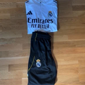 Real Madrid fotbollströja Adidas - Säljer en vit Real Madrid fotbollströja från Adidas med svart tryck och klubbmärke på bröstet. Tröjan har dragkedja vid halsen och svarta detaljer på axlarna. Materialet är lätt och andas, perfekt för träning eller match.