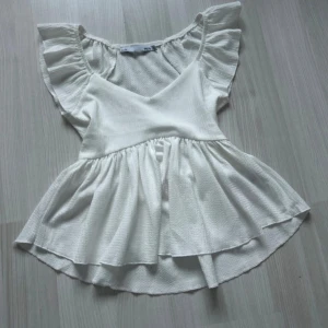 Vit volangblus från lager 157 XS - Supersöt vit blus från lager 157 i storlek XS. Toppen har v-ringning, volangärmar och en flowy, peplumliknande nederdel. Materialet är strukturerat och lätt, perfekt för varma dagar. Snygg att styla med jeans eller kjol för en fräsch look.