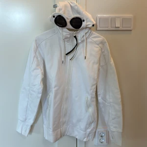 Vit CP Company goggle hoodie - Vit hoodiejacka från CP Company med ikoniska goggles i huvan. Jackan har dragkedja framtill, två fickor med dragkedja och ribbade muddar med tumhål. Snygg och clean design, perfekt för dig som gillar streetwear och vill sticka ut.