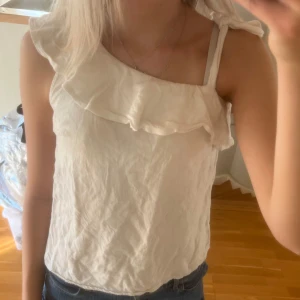 Vit omlottopp med volang och knyt - Säljer en söt vit topp i bomull med volang längs halsringningen och en axelbandslös design på ena sidan. Toppen har knyt på axeln och är ärmlös, perfekt för varma dagar. Enkel och stilren look som passar till jeans eller kjol. Litet håll som går och laga men som inte syns!