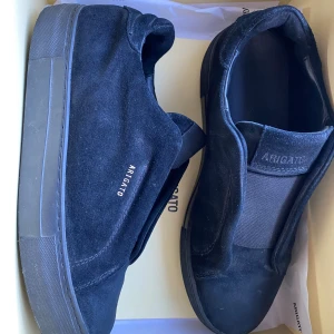 Svarta sneakers från Axel Arigato - Snygga svarta sneakers från Axel Arigato i mocka med diskret logga på sidan och platt sula. Skorna har en minimalistisk design med rund tå och elastisk panel för enkel påtagning. Perfekta för dig som gillar stilrena och moderna skor.