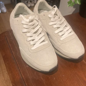 Beige Philippe Model sneakers i mocka - Snygga beige sneakers från Philippe Model, tillverkade i Italien. Skorna har en stilren design med rund tå, platt sula och snörning framtill. Utsidan är i mjuk mocka och insidan är fodrad i ljust material. Perfekta för dig som gillar minimalistisk och clean stil.