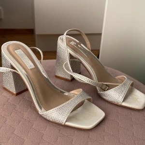 Glittriga sandaler med blockklack från Be Mine - Säljer ett par sandaler med bred blockklack och öppen tå från Be Mine. Skorna är täckta med glittriga stenar och har en elegant, ljus silvrig ton. Remmarna är tunna och satinklädda, och klacken är hög för extra statement. Perfekt för dig som vill sticka ut.