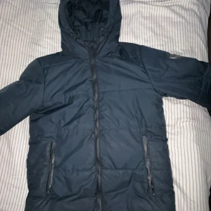 Mörkblå pufferjacka från EA7 Armani - Snygg mörkblå pufferjacka från EA7 Emporio Armani med huva och dragkedja framtill. Jackan har två fickor med dragkedja och en av fickorna med dragkedja funkar inte men det syns inte när man kollar på den. Och den har en stilren logga på ärmen. Perfekt för kalla dagar och ger en clean outfit. Har använt den jätte lite och är trött att ha den hemma nu. (Ny pris 1 200kr) pris kan diskuteras 