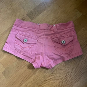 Rosa jeansshorts med fickor - Säljer ett par rosa jeansshorts med låg midja och coola fickdetaljer både fram och bak. Shortsen har bälteshällor, knappar och dragkedja framtill samt dekorativa sömmar. Perfekta för varma dagar och har en snygg, tight passform.