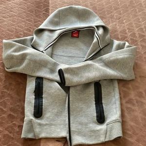 Grå NIKE TEECH FLEECE NY MODELL  - Snygg ljusgrå hoodie från Nike med svarta dragkedjor på fickorna och klassisk logga på bröstet. Tröjan har hel dragkedja, huva och är tillverkad i en mjuk bomulls- och polyestermix. Perfekt för chill eller träning.