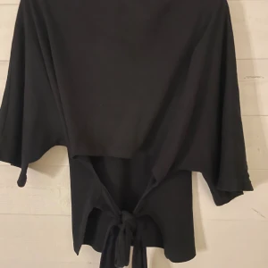 Svart knytblus från Nelly - Svart blus från Nelly med vida ärmar och snygg knytning framtill. Toppen har en lös passform och är tillverkad i mjukt material som känns skönt mot huden. Perfekt för dig som gillar stilrena och enkla plagg med en twist.