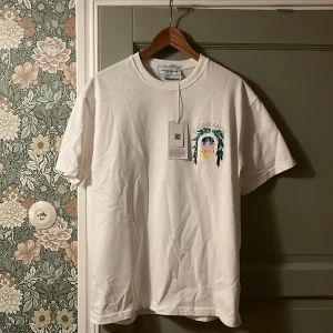 Vit t-shirt från Casablanca med tryck - Vit t-shirt från Casablanca med färgglatt tryck på bröstet som föreställer en byggnad i nattmiljö omgiven av gröna blad. Klassisk passform och rund halsringning. Mjuk bomullskvalitet och snygg detalj med logga ovanför motivet.