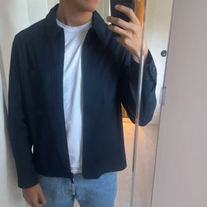 Marinblå overshirt från J.Lindeberg - Säljer en stilren marinblå overshirt från J.Lindeberg i bomullstwill. Jackan har klassisk krage, bröstficka och stängs med dragkedja. Perfekt för lager-på-lager och passar till jeans eller chinos för en clean look.    Sänker priset vid snabb affär