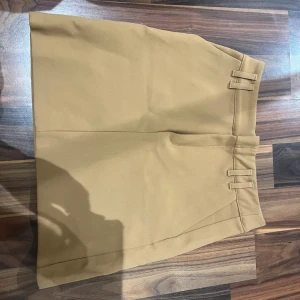 Beige kortkjol med bälteshällor - Snygg beige kortkjol med minimalistisk design och dubbla bälteshällor i midjan. Kjolen har en rak passform och är tillverkad i ett slätt tyg som ger en clean och stilren look. Perfekt för dig som gillar enkel och trendig stil.