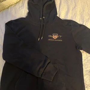 Mörkblå hoodie från GANT - Säljer en mörkblå hoodie från GANT med broderad logga och text på bröstet. Tröjan har huva med snörning, känguruficka och ribbade muddar. Perfekt för en chill och sportig stil.