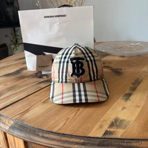Burberry keps  - Snygg keps från Burberry i deras ikoniska beigea rutmönster med svarta, vita och röda ränder. Framtill sitter ett broderat svart 'B'-emblem. Justerbar rem baktill med metallspänne. Perfekt accessoar för att lyfta din stil. Fint skick, köpt för 2999, vill bli av med den snabbt. Därav priset. Kom privat för mer information!