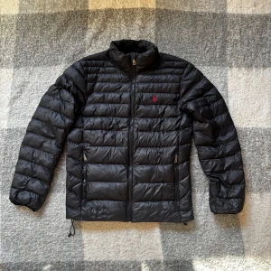 Ralph Lauren dunjacka – herr - Stilren svart dunjacka från Ralph Lauren i klassisk modell. Jackan har lätt fyllning som gör den både varm och smidig – perfekt för höst, vår och milda vinterdagar.  ✔️ Äkta Ralph Lauren ✔️ Svart med röd logga på bröstet ✔️ Två dragkedjefickor fram ✔️ Dragsko i nederkant för bättre passform  Skick: Väldigt fint begagnat skick, endast ett litet hål längst ner på jackan [se bild 5] Storlek: [xs]  Nypris ca 4199 kr.  Mitt pris: 2199 kr. (Prutbart)
