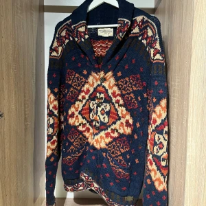 Mönstrad kofta från Ralph Lauren - kofta från Ralph Lauren Denim & Supply. Den är stickad i mörkblått med orange, rött och beige mönster, har stora knappar framtill och en bred krage. Pris satt ex frakt