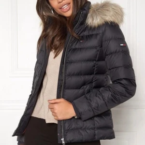 Svart dunjacka från Tommy Jeans - Snygg svart dunjacka från Tommy Jeans med fluffig fuskpäls på huvan. Jackan har quiltad design, dragkedja framtill och två fickor. Perfekt för kalla dagar och har en stilren look med diskret logga på bröstet och ärmen.
