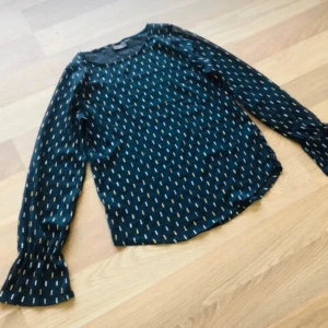 Mönstrad blus från ICHI med trumpetärm - Supersnygg blus från ICHI i mörkblått med små mönster i guld och vitt. Blusen har långa trumpetärmar och rund halsringning. Tunt, lätt material som ger en luftig känsla och cool look. Har aldrig används ny! Köptes för 300 kr!