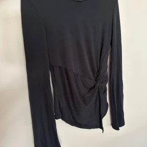 Svart långärmad topp från Gina Tricot - Svart långärmad topp från Gina Tricot i mjukt material. Toppen har en snygg knutdetalj i sidan som ger en cool twist till looken. Enkel och stilren design med rund halsringning, perfekt att matcha med jeans eller kjol.