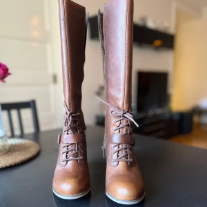 Bruna höga stövlar från Timberland - Snygga bruna stövlar från Timberland med högt skaft, snörning och dekorativt spänne. Tillverkade i skinn med grov sula och stabil klack. Insidan har ett mönstrat foder och dragkedja längs skaftet. #timberland