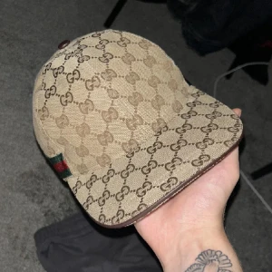 Beige keps med GG-mönster - Säljer en beige keps  med klassiskt GG-monogram över hela kepsen. Kepsen har bruna detaljer, justerbar rem bak i brunt läder och en grön-röd rand på sidan. Materialet är bomullscanvas och insidan är mörkbrun. Perfekt för dig som vill ha en lyxig streetstyle. Säljer så snabbt som möjligt vill bli av med den. 
