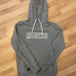 Grå hoodie från Morris med tryck - Grå hoodie från Morris med vit broderad logga på bröstet och vita snören i huvan. Klassisk känguruficka framtill och ribbade muddar. Mjuk och skön i bomullsblandning, perfekt för chill dagar.