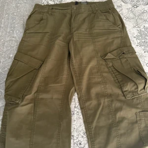 Olivgröna cargobyxor från H&M Divided - Säljer ett par olivgröna cargobyxor från H&M Divided i storlek 40. Byxorna har resår i midjan, stora sidofickor med lock och klassisk cargostil. Tillverkade i bomullsmix för en skön och avslappnad känsla. Perfekta för en chill och streetig look.
