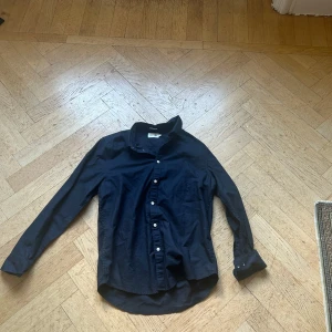 Skjorta från East West - Klassisk svart skjorta från East West i bomull. Skjortan har lång ärm, knappar framtill och en snygg krage. Perfekt för dig som gillar en stilren och enkel look. Passformen är normal och materialet känns mjukt mot huden.