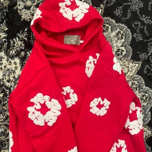 Denim Tears hoodie - Cool röd hoodie från Denim Tears med stora vita blommönster över hela tröjan. Tröjan har huva och en stor magficka framtill. Perfekt statement-plagg för dig som vill sticka ut med färg och mönster.