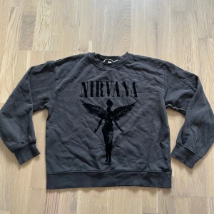 grå Nirvana sweatshirt - Cool grå/svart sweatshirt med Nirvana-tryck och ikonisk ängel på bröstet. Tröjan har rund hals, långa ärmar och ribbade muddar. Perfekt för dig som gillar bandmerch och vill ha en avslappnad stil.