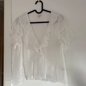 Volangblus från Nelly - Supersöt vit blus från Nelly med volanger längs ärmar och nederkant. Blusen har v-ringning, knappar framtill och är i ett lätt, luftigt material. (Använd endast två gånger). 