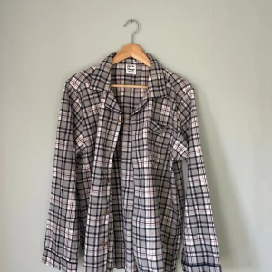 Rutig pyjamas skjorta från Time to Lounge M - Rutig skjorta i mjuk bomull från Time to Lounge. Skjortan har klassisk krage, knäppning framtill, bröstficka och långa ärmar. Färgerna är grå, vit, marinblå och röd. Perfekt för en chill och avslappnad stil.