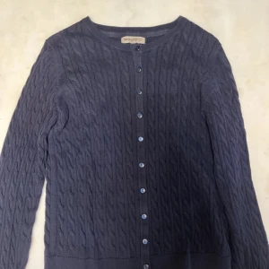 Mörkblå kabelstickad kofta Basic - Kabelstickad mörkblå kofta från Basic med rund halsringning och knappar hela vägen framtill. Långärmad modell med ribbade muddar och klassisk, stilren look. Perfekt att slänga över en t-shirt eller klänning för extra värme.