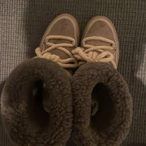 Bruna Inuikii moon boots med fårskinn - Säljer ett par bruna Inuikii moon boots med fluffigt fårskinnsfoder och grova beige snören. Skorna har en rund tå, platt sula och är perfekta för kalla dagar. Utsidan är i mocka och skinn, vilket ger en lyxig och mysig känsla.
