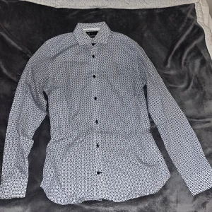 Vit och svart mönstrad skjorta Selected - Snygg vit skjorta från Selected Homme med svart prickigt mönster. Skjortan har klassisk krage, svarta knappar och lång ärm. Perfekt för dig som gillar stilrena detaljer och vill sticka ut lite extra.