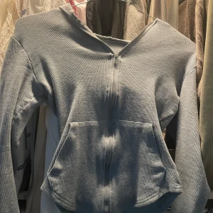 Grå ribbad kofta med dragkedja - Grå kofta med ribbad struktur, hel dragkedja framtill och två små fickor. Tröjan har huva med snörning och långa ärmar.Den sitter väldigt tajt vid midjan och är kort