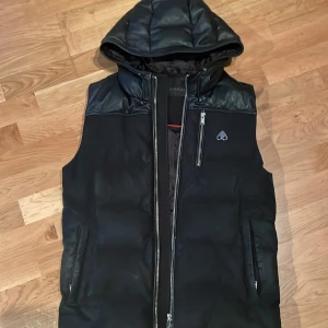 Svart dunväst från Moose Knuckles - Svart dunväst från Moose Knuckles (Storlek S men passar M) med huva och detaljer i skinnimitation över axlar och huva. Västen har två sidofickor med dragkedja, en bröstficka med dragkedja och logga på bröstet. Stängs med en kraftig dragkedja framtill.