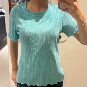 Turkos ribbad topp Silvio Viselle - Turkos ribbad topp från Silvio Viselle med korta ärmar och coola metallöljetter med snörningsdetalj vid halsen. Den har ett glansigt material. 