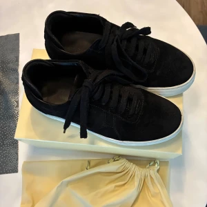 Axel Arigato svarta platform sneakers - Svarta sneakers från Axel Arigato med stilren design och vit platt sula. Skorna är tillverkade i mocka och har snörning framtill. Diskret guldfärgad logga på sidan ger en exklusiv touch. Perfekta för dig som gillar minimalistisk och modern stil.