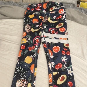 Färgglada leggings från Stronger XS - Säljer ett par leggings från Stronger i storlek XS med färgglatt fruktmönster och vita ränder på benen. Byxorna är mörkblå i grunden och har inslag av avokado, apelsin, körsbär och blommor. Materialet är stretchigt i polyester och elastan, perfekt för träning.