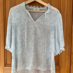 Ljusblå blus med volang - Supersöt ljusblå blus med vitt blommigt mönster 🌸 blusen har v-ringning med knytband, volangdetaljer och trekvartsärmar 💕nyskick men säljer billigt!!