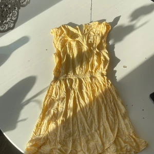 Gul kort sommarklänning med volang - Söt gul kortklänning med små vita blommor och volangdetaljer. Klänningen har resår i midjan, är ärmlös och har en lätt omlottkänsla upptill. Perfekt för soliga dagar och ger en fräsch och glad look.