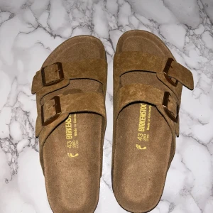 Bruna mockasandaler från Birkenstock - Säljer ett par klassiska sandaler från Birkenstock i brun mocka med två breda remmar och justerbara metallspännen. Skorna har en anatomiskt utformad fotbädd och svart sula med mönster. Perfekta för chill dagar och enkel stil.