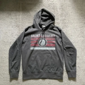 Grå Saint Laurent hoodie med tryck - Säljer en grå hoodie från Saint Laurent med huva och snörning. Framsidan har ett coolt tryck med röda och vita ränder samt SL-emblem. Klassisk känguruficka och ribbade muddar. Perfekt för dig som gillar streetstyle och designerplagg. Står M men passar mer S.