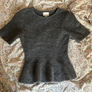 Grå stickad topp med volangkant - Mysig grå stickad topp med korta ärmar och rund halsringning. Toppen har en figurnära passform och en snygg volangkant nedtill som ger en feminin siluett. Perfekt att styla med jeans eller kjol för en trendig look.