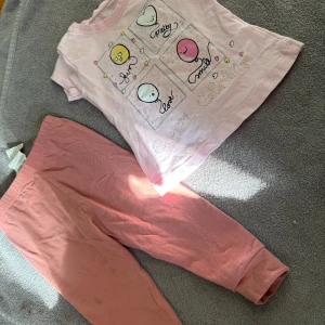 Rosa t-shirt med ballongtryck från Peri Masali - Söt ljusrosa t-shirt från Peri Masali med korta ärmar och gulligt tryck av ballonger och hjärtan i vitt, gult och rosa. Texten 'fun', 'pretty', 'love', 'smile' och små pärldetaljer ger extra charm. Perfekt för små barn som gillar färg och lekfulla motiv.